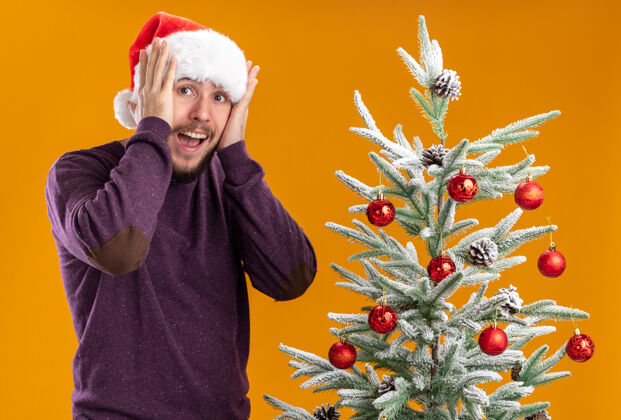 站着穿着紫色毛衣 戴着圣诞帽的年轻人站在橙色背景下的圣诞树旁 看上去既惊讶又惊讶圣诞老人惊讶帽子