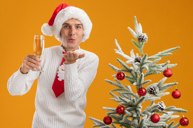 高兴高兴的年轻帅哥戴着圣诞帽 打着圣诞老人的领带 站在装饰好的圣诞树旁 手里拿着一杯香槟 看着摄像机 在橙色的背景下 孤零零地送上一吻吹装饰领带