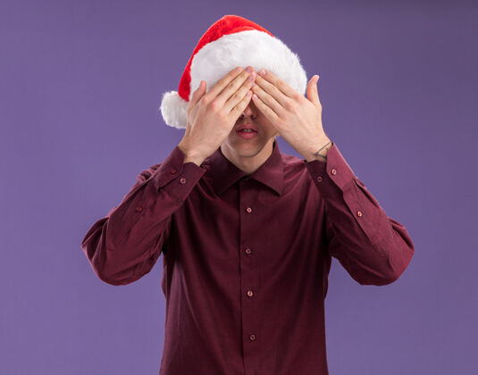 圣诞老人年轻的金发男子戴着圣诞帽 戴着一副眼镜 双手孤立在紫色背景上遮住眼睛眼睛年轻眼镜