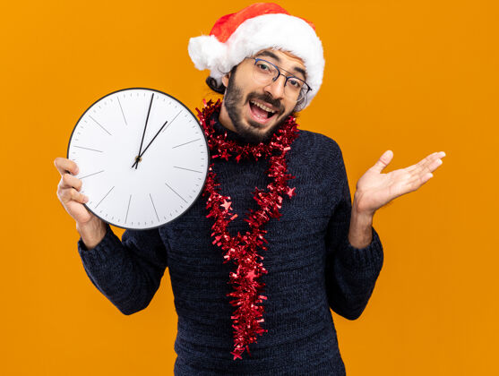 抱着高兴的年轻帅哥戴着圣诞帽 脖子上戴着花环 手里拿着挂在橙色背景上的挂钟帽子圣诞钟