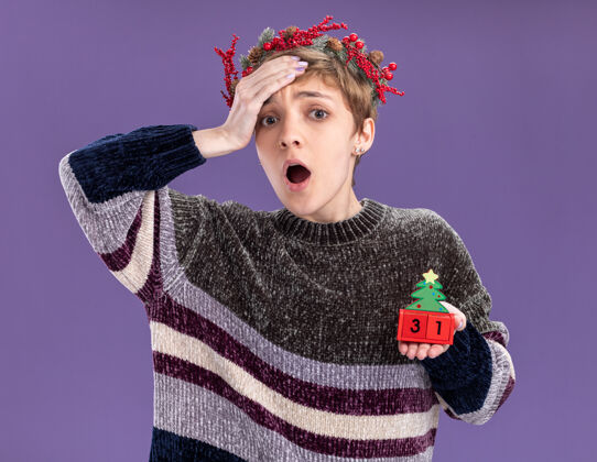 快乐关心的年轻漂亮女孩戴着圣诞花环拿着圣诞树玩具和日期看着相机手放在额头上隔离在紫色背景上手圣诞额头