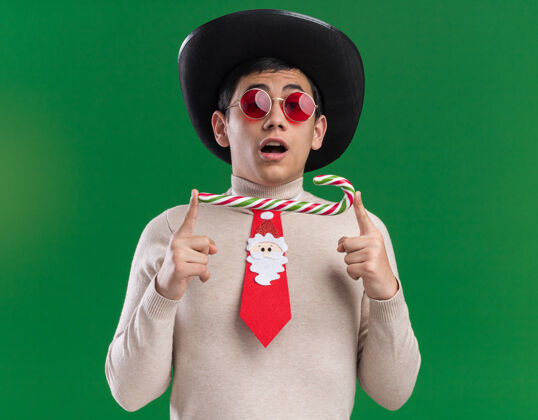 糖果惊讶的年轻人戴着帽子 打着圣诞领带 戴着眼镜 手里拿着圣诞糖果 背景是绿色的小伙子圣诞节帽子
