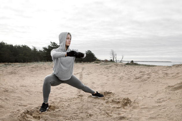 年轻人穿着手套 连帽衫和运动鞋的时尚年轻女运动员做站立瑜伽姿势 强健双腿 为有氧运动准备肌肉自信的女运动员在沙滩上锻炼漂亮运动锻炼