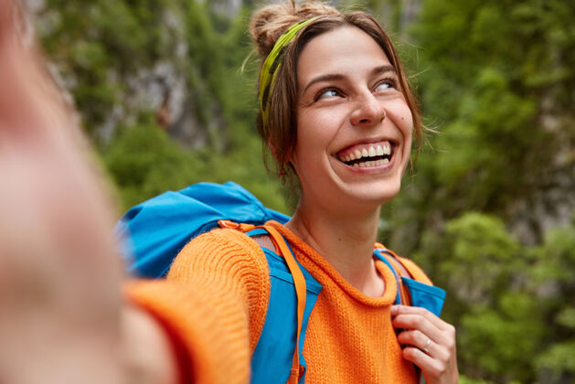 能量开朗的女探险家在绿林中有着美妙的旅程 伸出手自拍 背着背包站在户外 笑容灿烂 专注在一旁大家好人类自拍白天