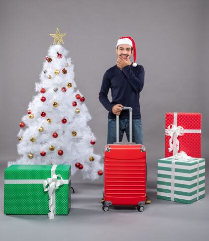 微笑微笑的圣诞老人拿着红色的旅行箱 站在白色圣诞树旁 圣诞玩具五颜六色盒子男人旅行袋