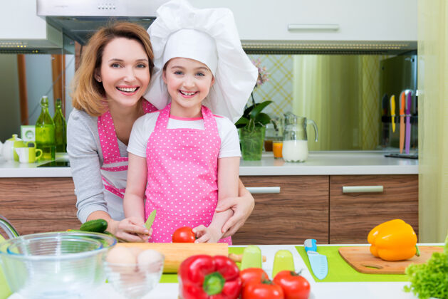 健康快乐的年轻母亲和穿着粉色围裙的女儿在厨房做饭的画像帽子孩子餐