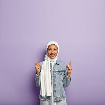 垂直开朗女性的垂直镜头指向食指上方 有愉快的微笑 显示空白 按照传统戴着白色面纱 隔离在紫色的墙壁上有自由的空间个性伊斯兰教指向