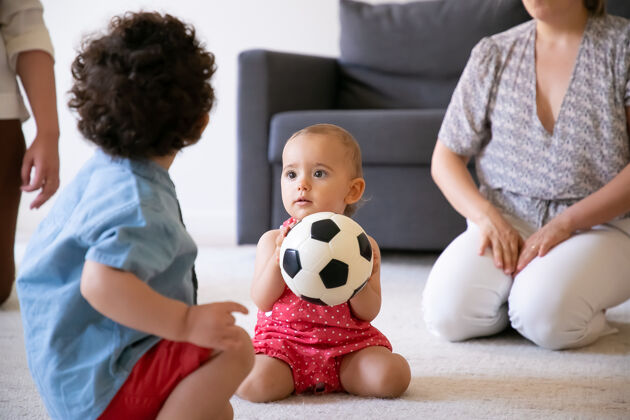 视图有趣的女婴拿着足球 坐在地毯上和弟弟在房间里玩不规则的妈妈和孩子们玩得很开心卷发男孩的后视图室内家庭 周末和童年的概念生活周末女人