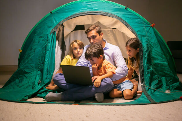 电影可爱的孩子们和爸爸一起在家里的帐篷里放松 在笔记本电脑上看电影可爱的孩子和中年爸爸坐在一起玩童年 家庭时间和周末的概念姐姐童年玩耍