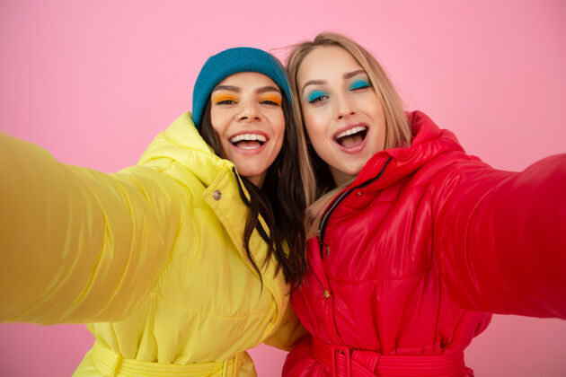 羽绒服两个迷人的女人穿着鲜艳的红黄相间的冬季羽绒服 在粉色的背景下摆着姿势 朋友们在一起玩得很开心 温暖的服装时尚潮流 自拍外套女孩衣服