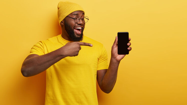 暗哑浓密鬃毛的黑人男子 指着智能手机设备 在空白屏幕上显示您的促销内容 戴着头饰和休闲黄色t恤 为客户宣传新设备短信手机成人