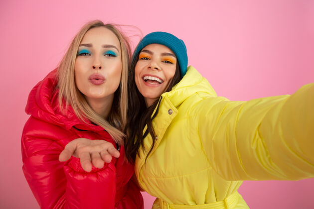 年轻两个迷人的女人穿着鲜艳的红黄相间的冬季羽绒服 在粉色的背景下摆着姿势 朋友们在一起玩得很开心 温暖的服装时尚潮流 自拍温暖的衣服套装五颜六色