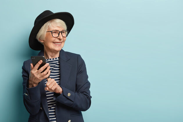 年龄老美女在手机上输入短信 使用新的在线应用程序 有积极的面部表情 目光移开 戴着时髦的头饰和外套 站在蓝色的墙上 自由的空间时尚眼镜牙齿