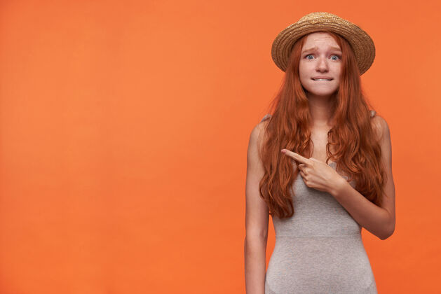 自然一张室内照片 困惑的年轻红发女子 卷曲的长发 穿着灰色衬衫 戴着草帽 孤立在橙色背景下 一边用食指 一边看着相机 一边咬着屁股食指发型提高