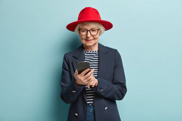 浏览有魅力的老妇人的照片等待反馈 集中在手机上 是真正的购物狂 戴着红帽子和正式的衣服 隔着蓝色的墙壁 阅读收到的信息聊天外套夹克