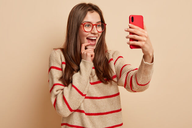 摄影快乐靓女拍搞笑照片 在现代手机上点击自拍照片 在社交网络上发帖 喜欢自己拍照 戴透明眼镜 穿休闲套头衫 与世隔绝迷人视频通话眼镜