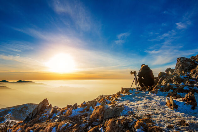日落日落时分 专业摄影师用相机在岩石峰的三脚架上拍照摄影师徒步旅行男孩