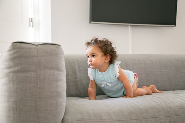 室内严肃可爱的深色卷发女婴穿着淡蓝色的布 爬在家里的沙发上 望着别处孩子在家和童年的概念混合混合赛车头发