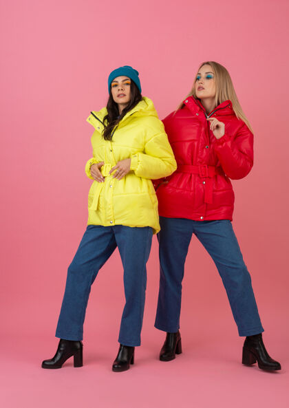 蓝色两个迷人的女孩在粉红色的背景下 穿着鲜艳的红色和黄色的彩色冬季羽绒服摆姿势彩色保暖女士