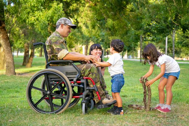 孩子快乐兴奋的妈妈和残疾军人爸爸坐在轮椅上和孩子们一起在户外消遣 在草地上放柴火残疾退伍军人或家庭户外概念支持残疾人观看
