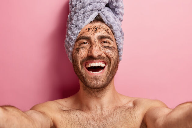 健康健康积极的男人自拍 用咖啡擦洗面部皮肤 有清洁程序 赤裸上身在粉色背景下摆姿势 头上放毛巾美容治疗高兴男性