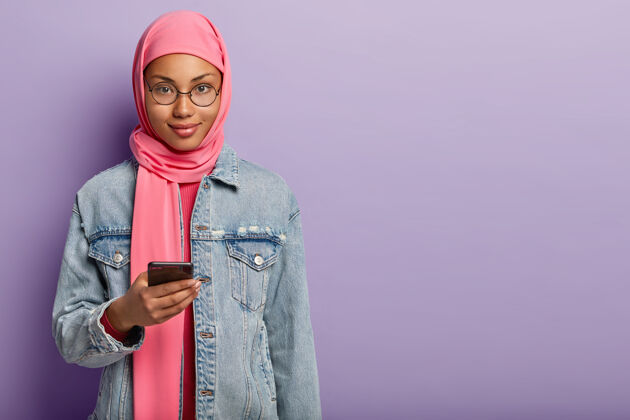 冲浪帅气的黑皮肤女人镜头穿着粉色头巾和牛仔外套 手持现代手机 等待重要电话 站在空白的紫色墙壁上肖像短信头巾