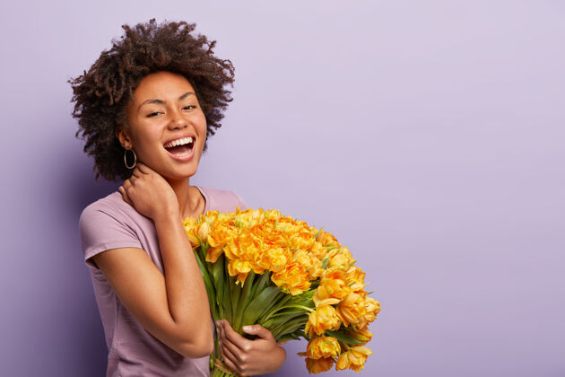 积极快乐的黑皮肤女人的侧拍欢笑 触摸脖子 拿着黄色的郁金香 穿着紫色的t恤 收到鲜花和赞美的喜悦 在紫色的墙上摆姿势 自由的空间欣喜若狂高兴清爽