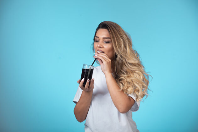 成人穿着白衬衫的年轻女孩拿着一杯黑鸡尾酒 在烟斗上检查味道摆姿势人类果汁
