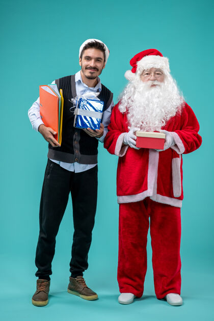 圣诞快乐圣诞老人与年轻男性和礼物在蓝色墙上的正面视图前面庆祝假期