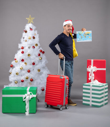 购物带着黄色背包的年轻人拿着圣诞树旁的地图 礼物是灰色的年轻人圣诞树圣诞帽