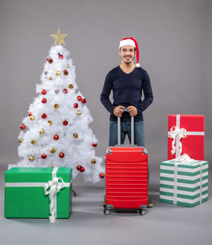 微笑圣诞树旁 微笑的圣诞老人手里拿着红色的旅行箱圣诞帽包庆祝