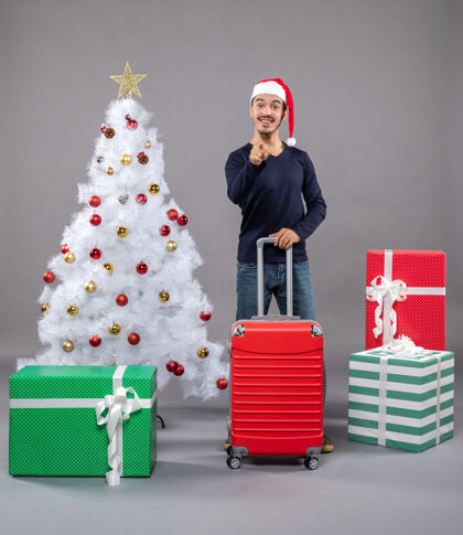 圣诞树圣诞老人戴着圣诞帽 拿着红色手提箱 在白色圣诞树旁放着五颜六色的圣诞玩具礼物举行旅行袋