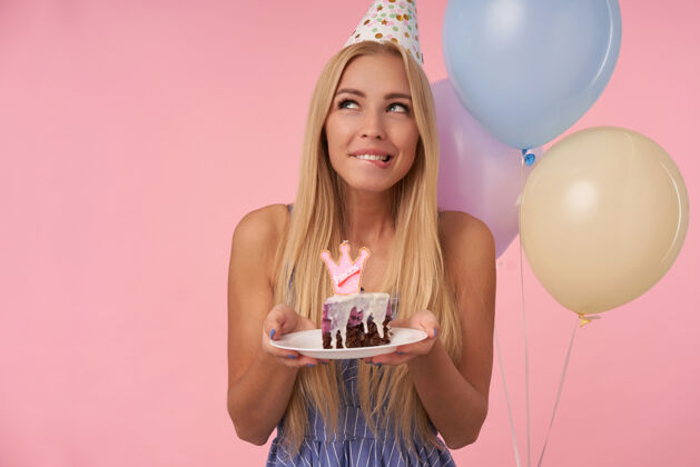 节日室内拍摄迷人的长发金发女士在五颜六色的气球里欢呼雀跃 拿着蛋糕 若有所思地看着一边 咬着粉色背景下的垫子发型裙子室内