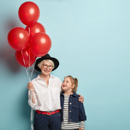 积极纵向形象：高兴的奶奶和小女孩儿拥抱 有美好的关系 一起庆祝节日 手持红气球 享受生日聚会 孤立的蓝色家庭肖像积极咯咯笑老人