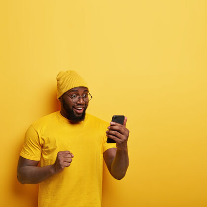 头饰喜出望外的非洲男人 浓密的头发 高兴地看着手机 心情乐观 庆祝好消息 握紧拳头 戴着黄色时髦的帽子眼镜手机短信