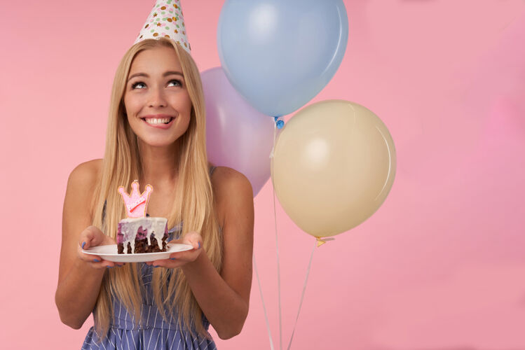 金发梦幻般的金发美女拿着蜡烛拿着一块蛋糕 开心地笑着 在粉色背景上摆着一堆五颜六色的氦气球饮食空气20多岁