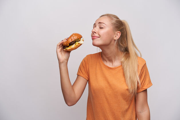 情绪摄影棚拍摄了一个积极的金发女郎 扎着马尾辫 手上拿着汉堡包 非常愉快地品尝着美味的饭菜 站在白色的背景下T恤美味发型