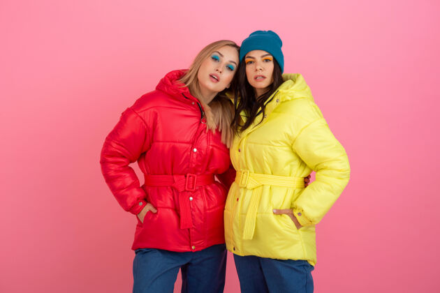羽绒服两个迷人的女孩在粉红色的背景下 穿着鲜艳的红色和黄色的彩色冬季羽绒服摆姿势保暖衣服女士外套