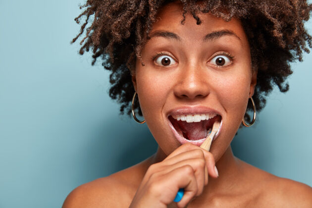 惊讶牙齿卫生和牙科概念头像是一位年轻的非洲裔美国妇女 头发蓬松 用牙刷和牙膏刷牙 眼睛被 眼睛被蓝色隔离卷发洗印象深刻