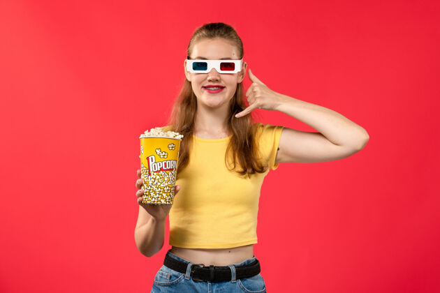 年轻的前视图年轻的女性在电影院拿着爆米花包戴着d太阳镜在红墙电影院看女性趣味电影电影爆米花电影