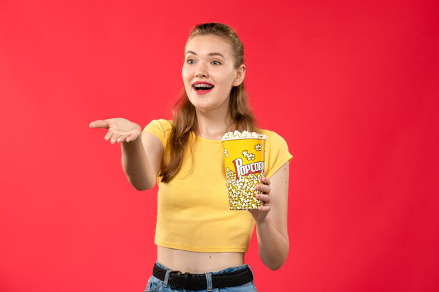 漂亮正面图年轻女性在电影院拿着爆米花包在浅红的墙上看电影剧院成人年轻