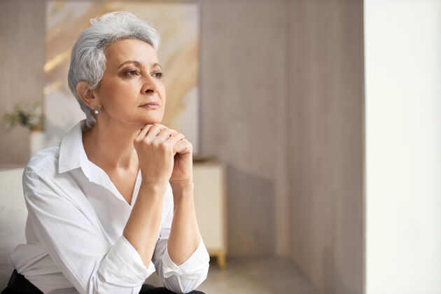 室内严肃优雅的退休妇女 短发 手放在下巴下 在室内摆姿势 带着沉思的表情看着别处 思考着一些想法或决定体贴沉思思考