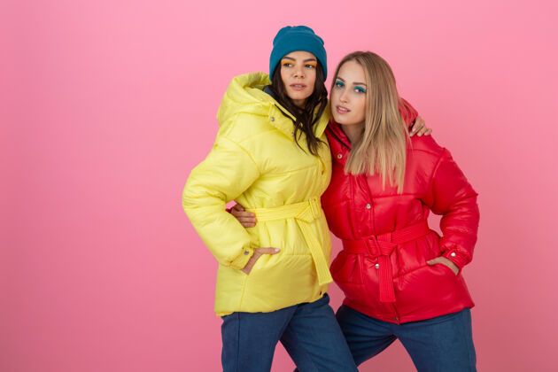 女士两个迷人的女孩在粉红色的背景下 穿着鲜艳的红色和黄色的彩色冬季羽绒服摆姿势女孩保暖时尚