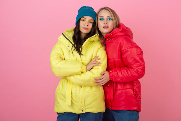 时尚两个迷人的女孩在粉红色的背景下 穿着鲜艳的红色和黄色的彩色冬季羽绒服摆姿势羽绒服彩色休闲