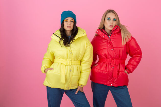 黄色两个迷人的女孩在粉红色的背景下 穿着鲜艳的红色和黄色的彩色冬季羽绒服摆姿势保暖时尚年轻