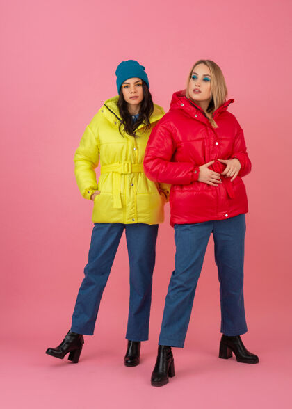 套装两个迷人的女孩在粉红色的背景下 穿着鲜艳的红色和黄色的彩色冬季羽绒服摆姿势保暖衣服衣服时尚