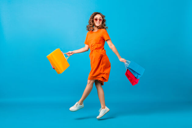 女孩迷人的快乐微笑时尚女性购物狂穿着橙色时髦的超大号连衣裙在蓝色工作室背景上拿着购物袋跳跃女女士零售