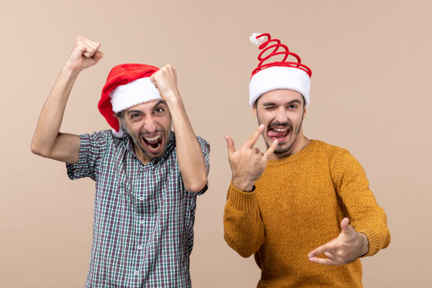 帽子两个戴着圣诞帽的兴高采烈的家伙在米色孤立的背景下展示他们的幸福圣诞老人前面米色