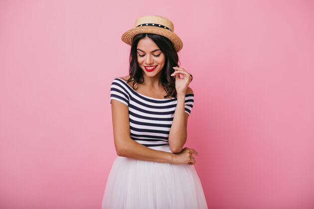 休闲带着草帽的沉思的笑女孩在拍照时俯视着穿着白裙子的浪漫的黑发女士的肖像笑室内粉色