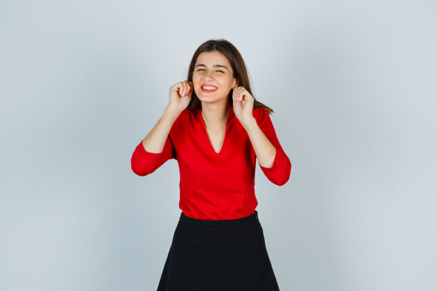 快乐穿着红衬衫的年轻女士 裙子拉着耳垂 看起来很滑稽衬衫有趣表情